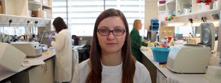 Picture of Gosia smiling in her lab coat, with scientists working behind her in the lab.
