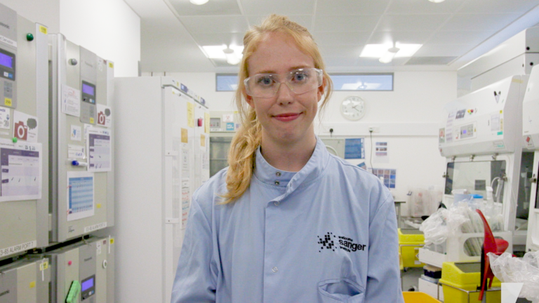 Picture of Lucy smiling, she is in a lab coat and protective glasses in the lab.