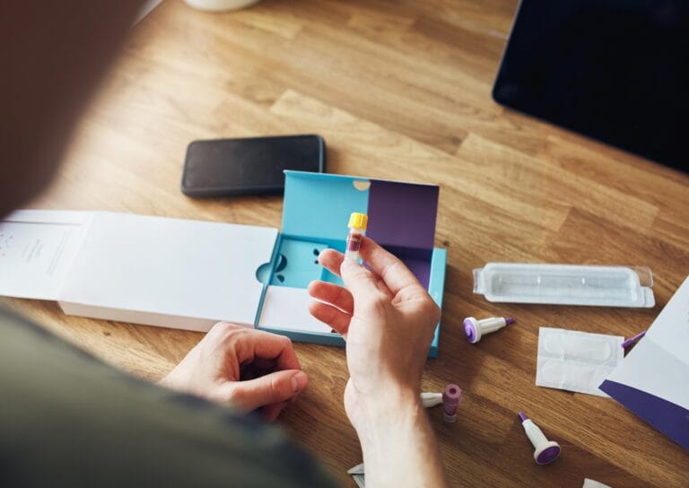 Picture of a male holding a genetic testing kit in his hands, on the desk below the rest of the kit is laid out.
