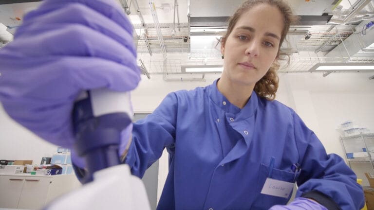A picture of Louise Aigrain, a senior staff scientist. Louise is wearing a lab coat with gloves on, she is conducting a science experiment.