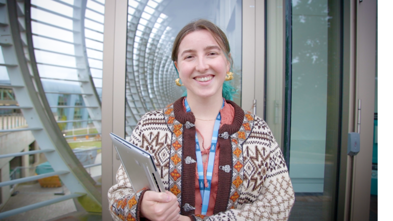 Picture of Jess smiling, holding her laptop on the Wellcome Genome Campus.