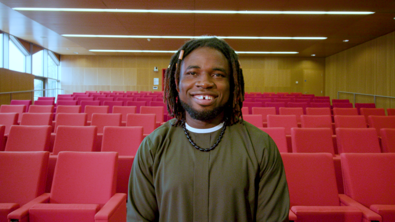 Picture of Joshua smiling, standing in front of a large theatre with multiple red empty chairs behind him. Joshua is a Product Manager for Wellcome Sanger Institute.