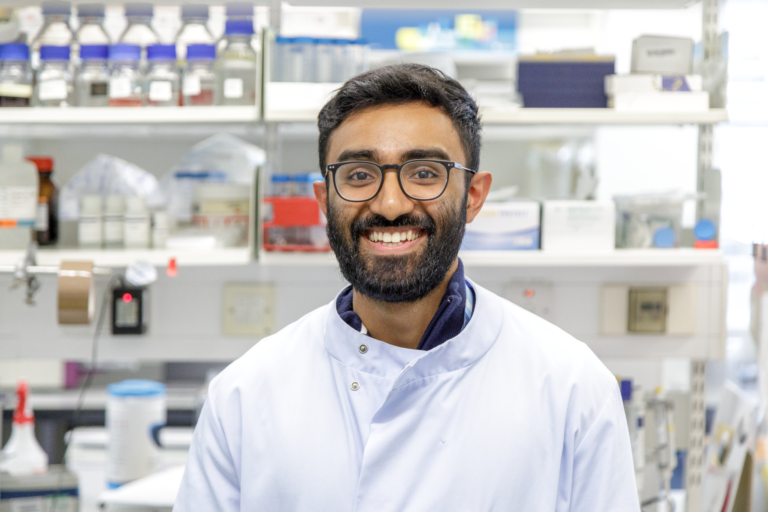 Picture of Abey smiling brightly in his lab coat. Abey is standing in a lab office space.