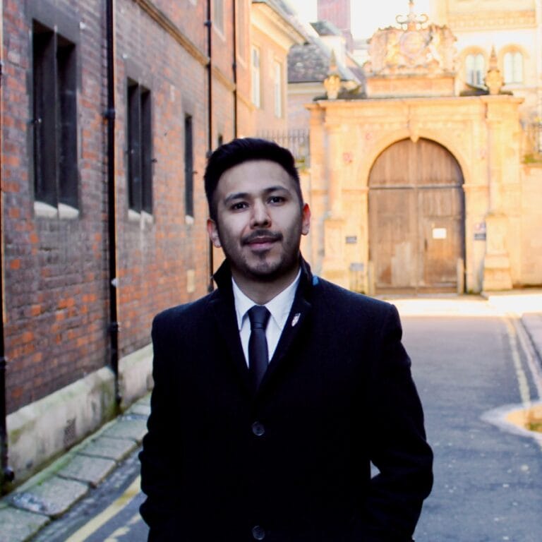 Image of Adrian, a man wearing a suit and tie in front of some red bricked buildings. Image of Adrian, a man wearing a suit and tie in front of some red bricked buildings.