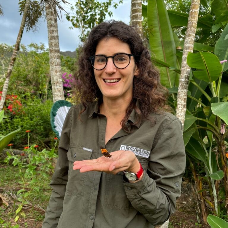 Image of Joana, a woman out in the jungle wearing a green top, holding a colourful butterfly in her hand. Image of Joana, a woman out in the jungle wearing a green top, holding a colourful butterfly in her hand.