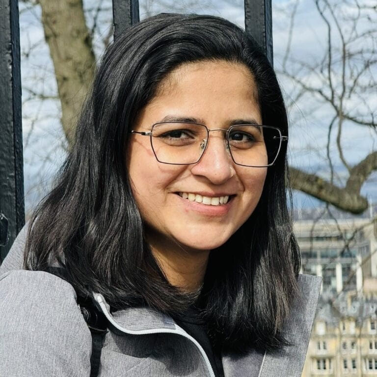Image of Nisha, a woman wearing glasses, outside, smiling. Image of Nisha, a woman wearing glasses, outside, smiling.